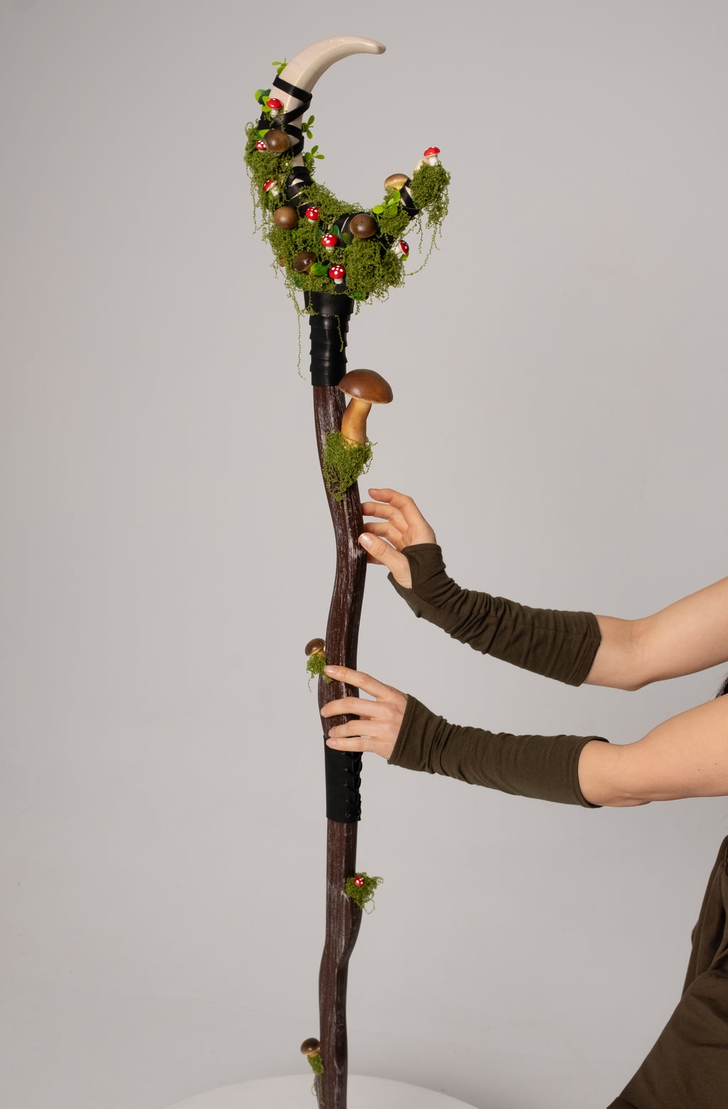 Decorative staff with greenery and small figures held by a person wearing brown sleeves against a gray background