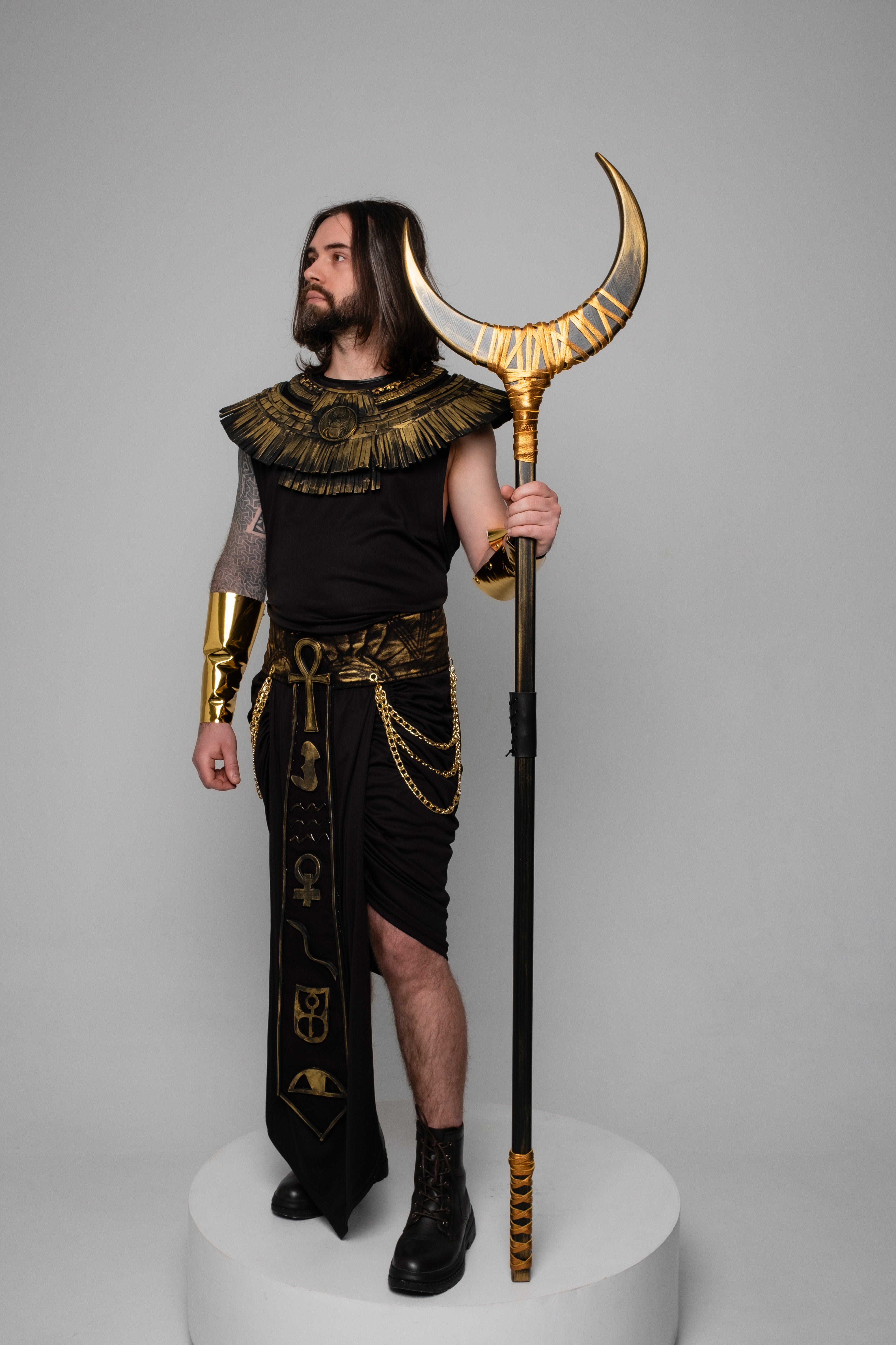 Person in Egyptian-themed costume holding a staff with a crescent moon design on a gray background