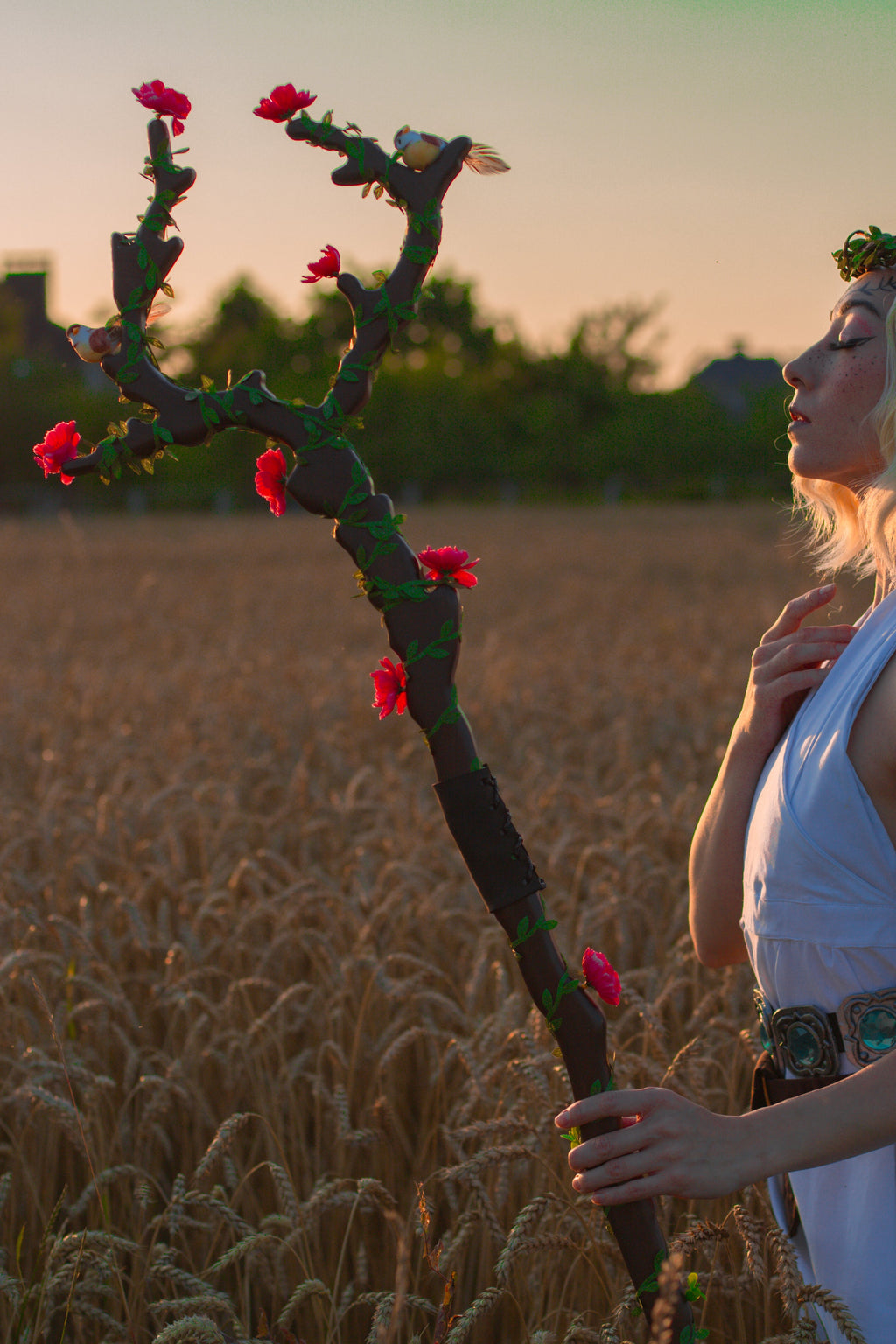 Woman in a white dress holding a staff with flowers in a wheat field at sunset