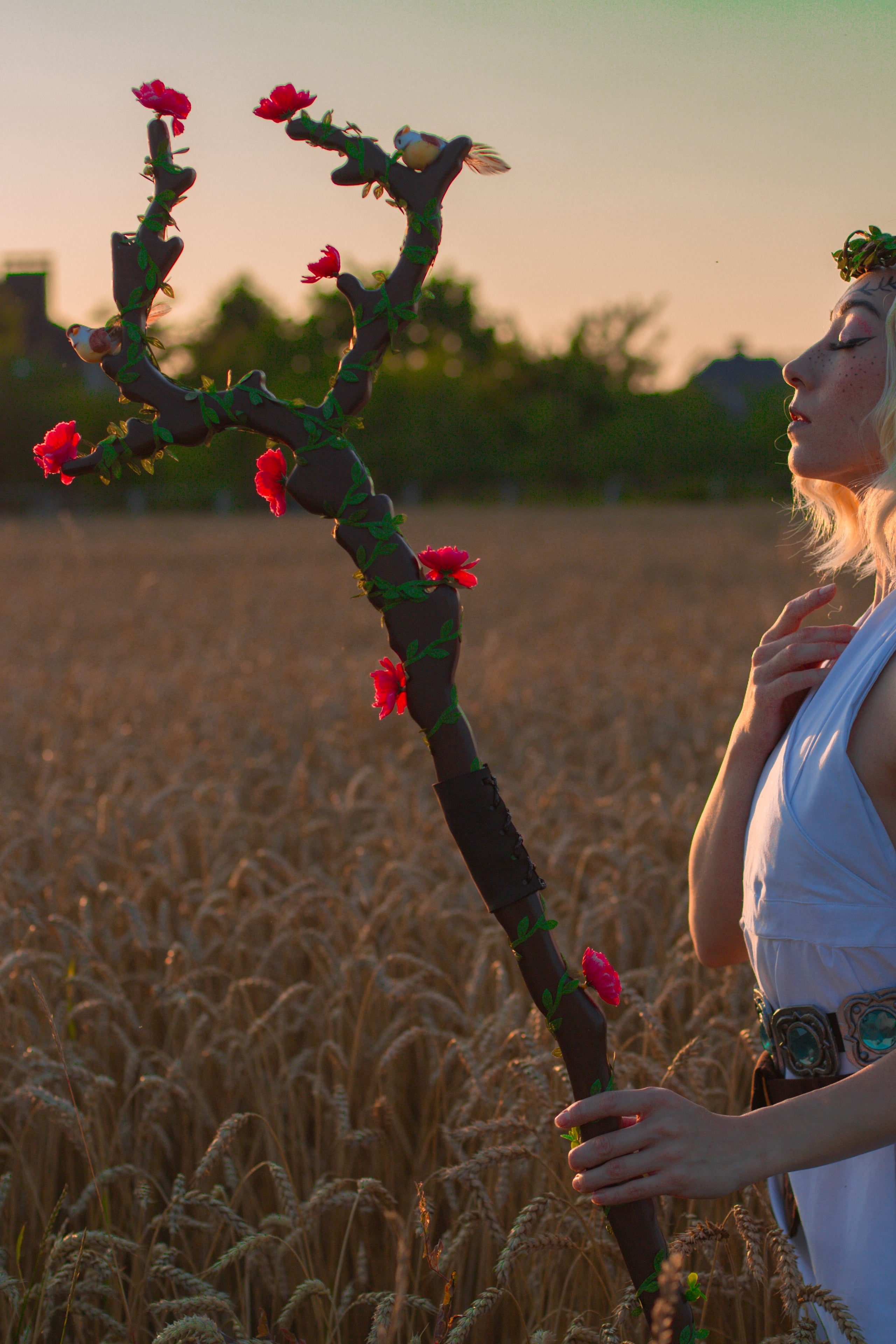 Woman in a white dress holding a staff with flowers in a wheat field at sunset