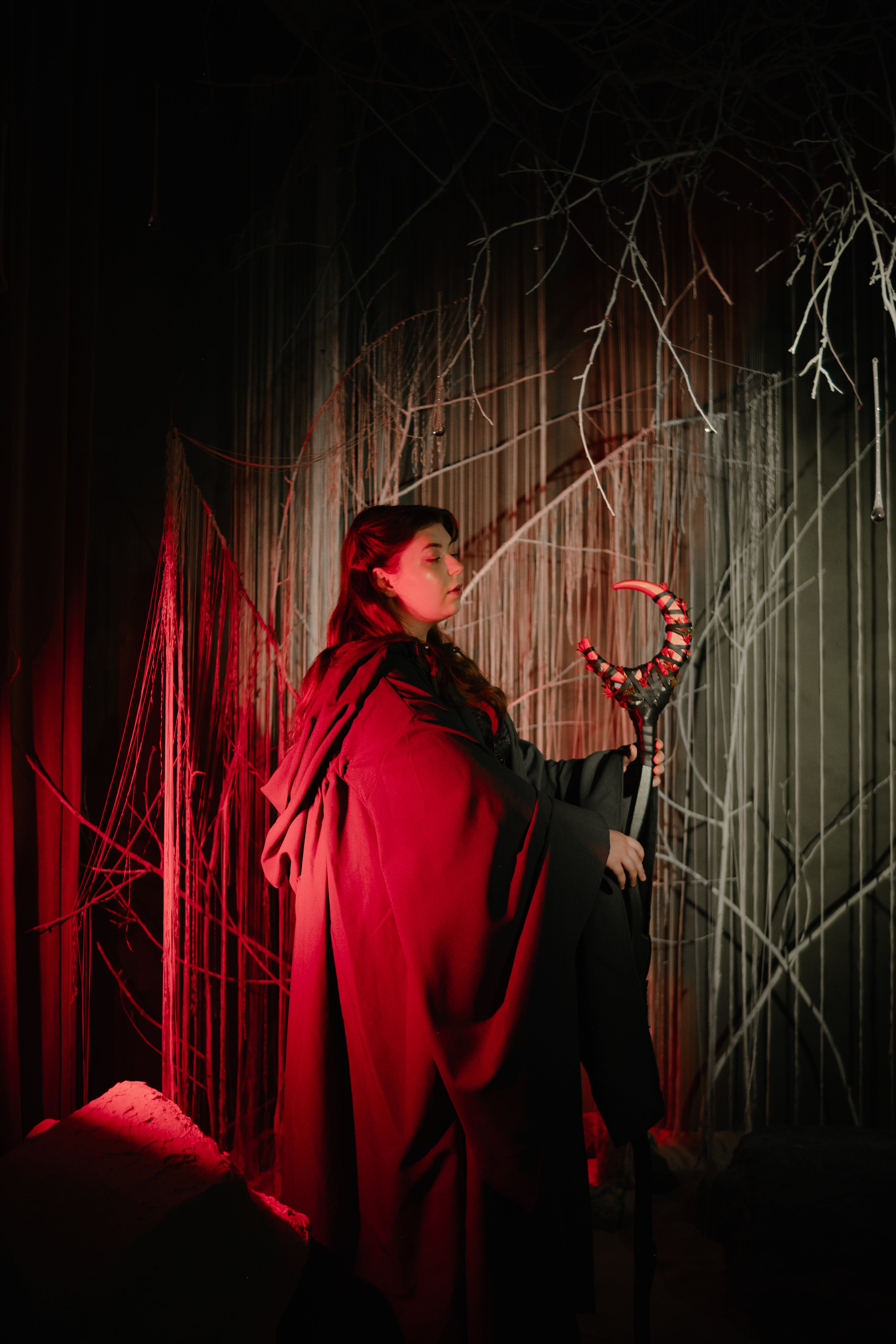 Person in a red cloak holding a staff with a glowing red tip against a dark background with abstract white lines.