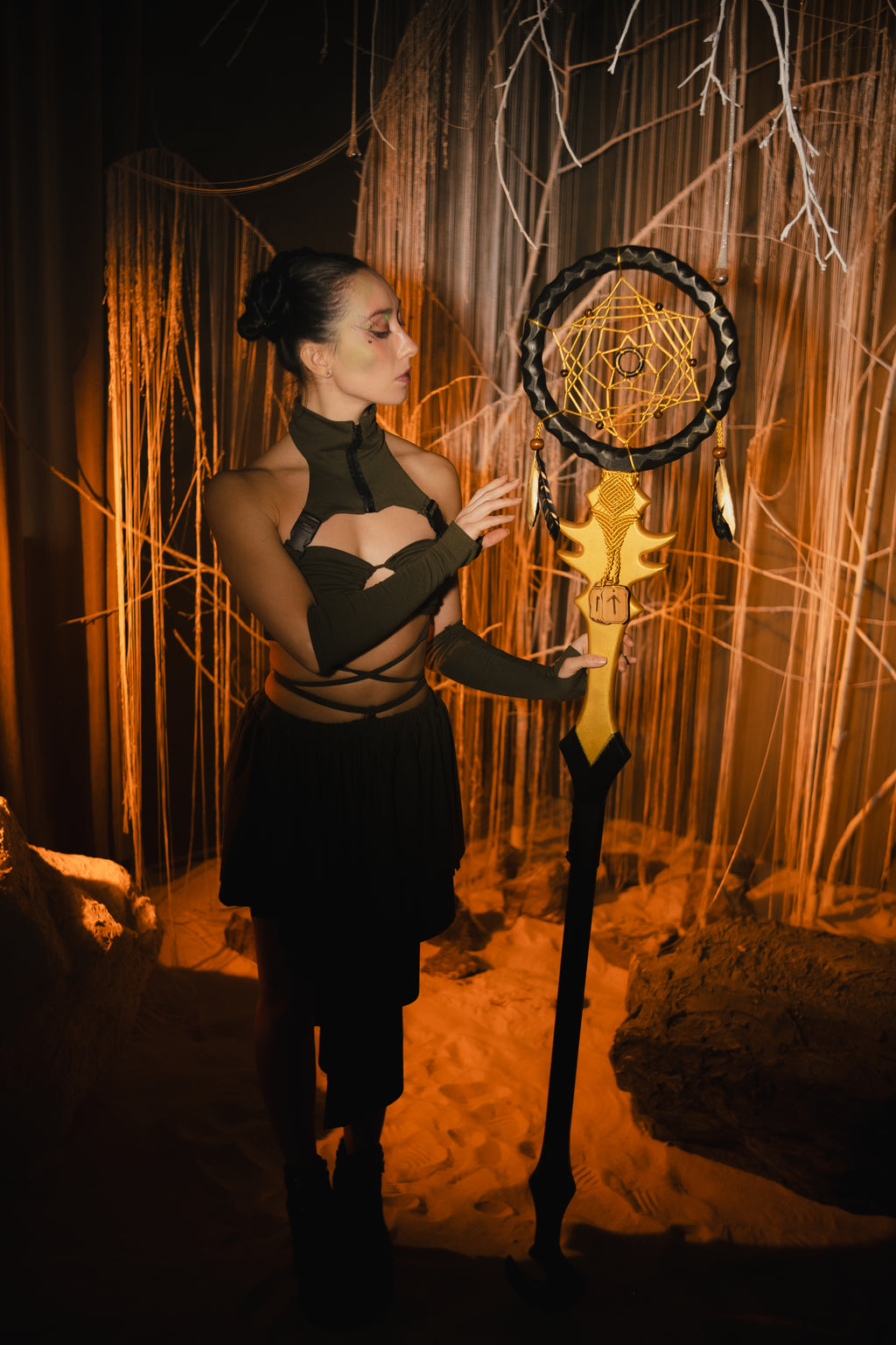Woman holding a large decorative staff with a dreamcatcher against a textured wall.