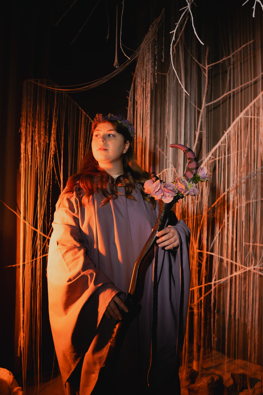 Person in a costume holding a floral staff against a dark, textured backdrop