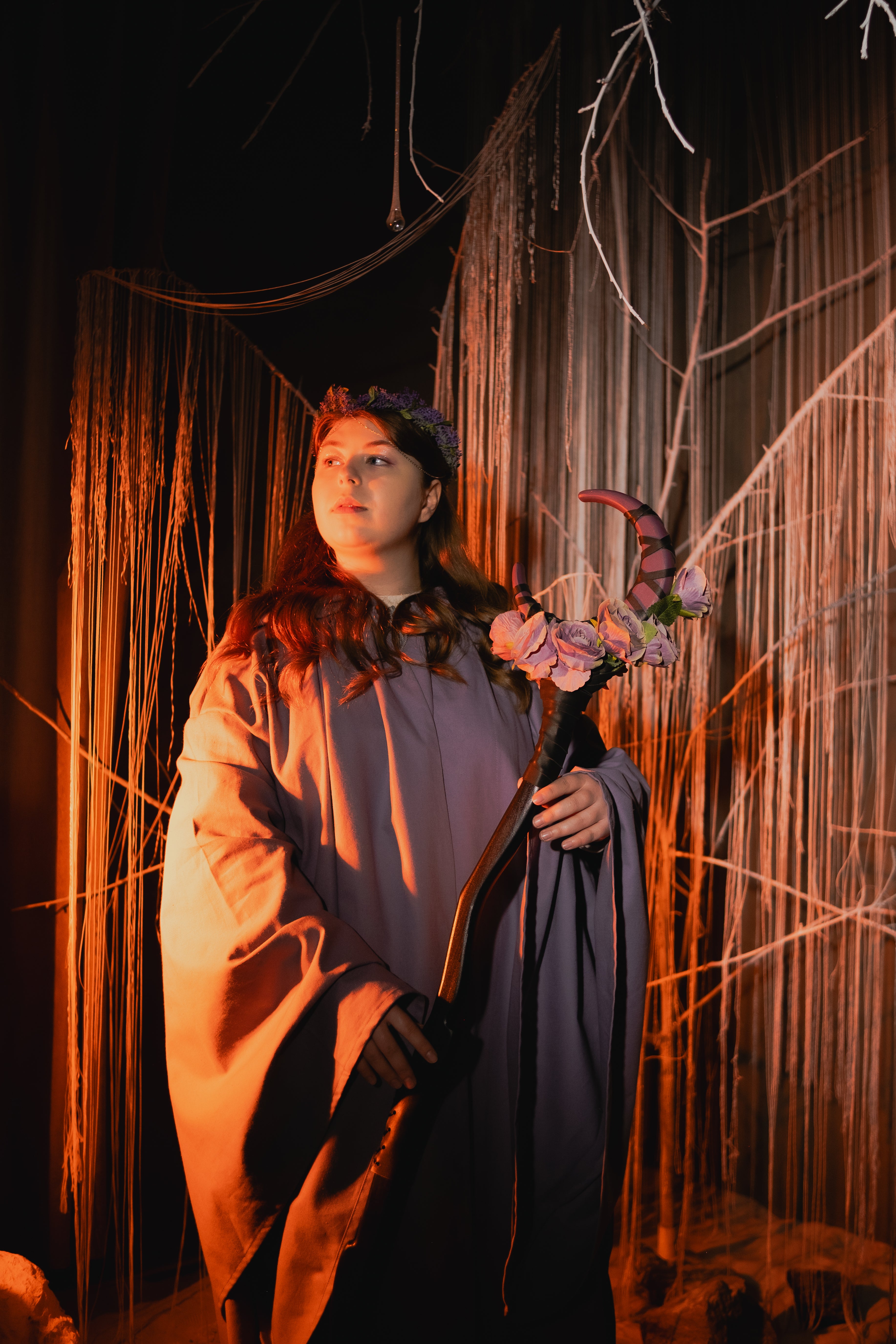 Person in a costume holding a floral staff against a dark, textured backdrop