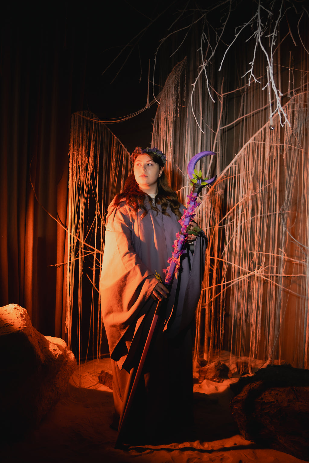 Person in a costume holding a purple staff against a dark background with draped fabric and branches.