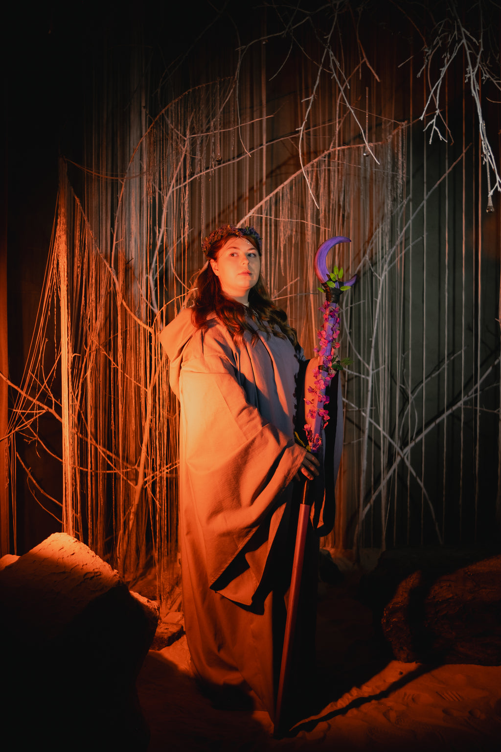 Person in a long coat holding a staff with a purple moon symbol, standing against a dark background with string art.